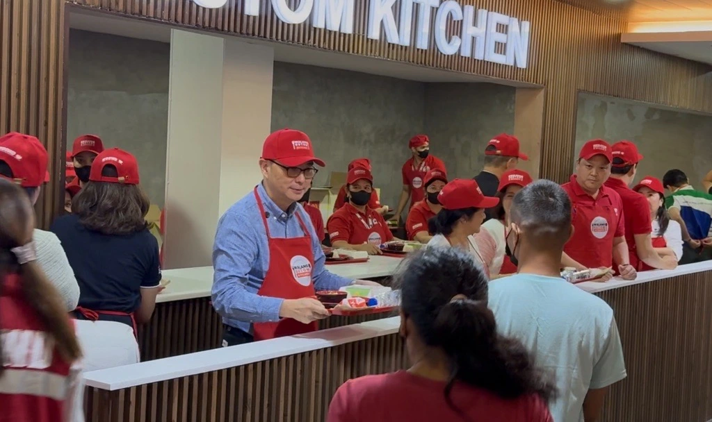 Staff in red caps and aprons serve meals at a long counter in a cafeteria setting under a 'KITCHEN' sign.