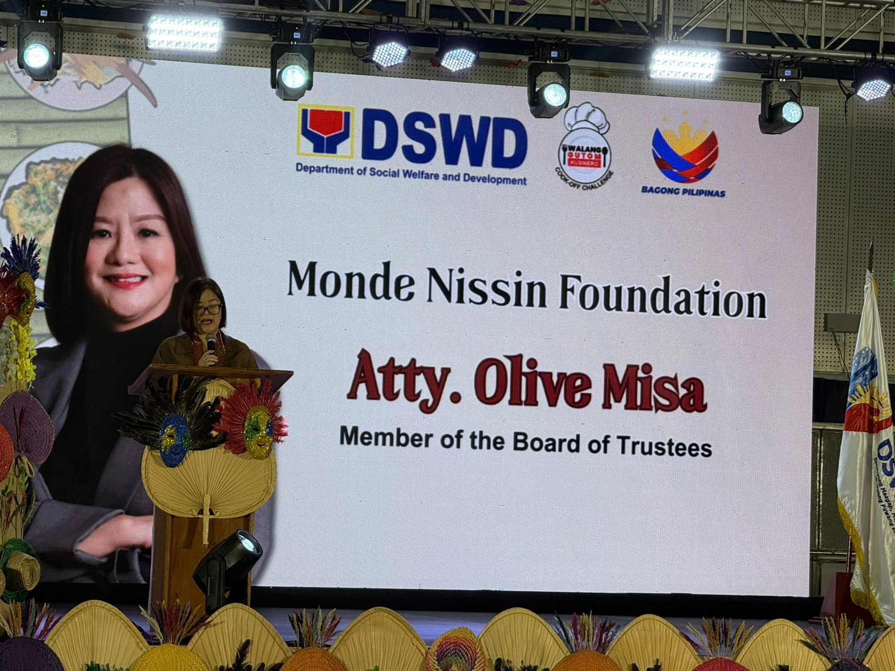 Speaker at a podium delivering remarks on a stage with a large screen showing Monde Nissin Foundation and DSWD logos. Decorative fans line the front of the stage.