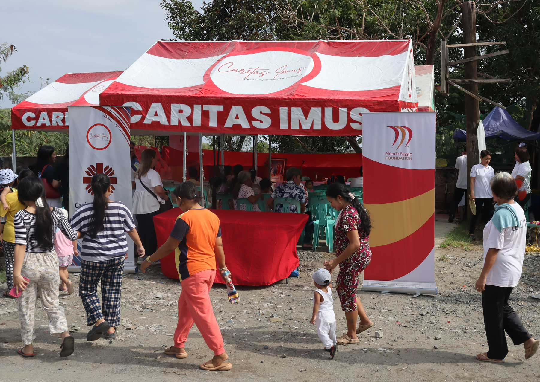 Outdoor charity event under a red-and-white canopy with people gathering at a table booth and banners.