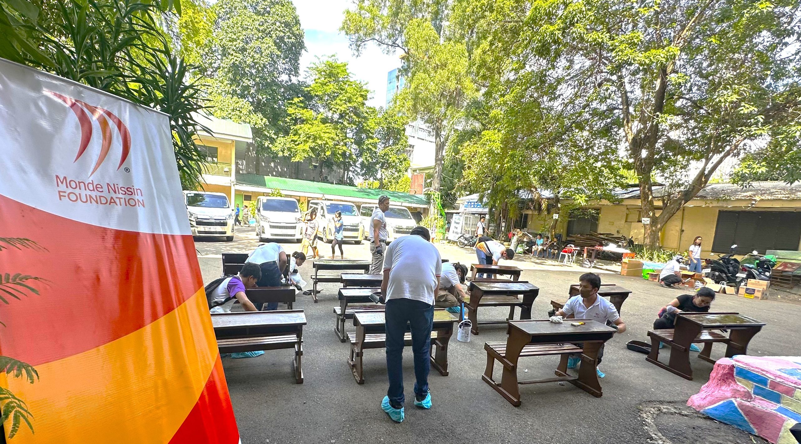 Outdoor community event under a Monde Nissin Foundation banner; people sit at benches setting up or receiving services in a sunny parking lot surrounded by trees.